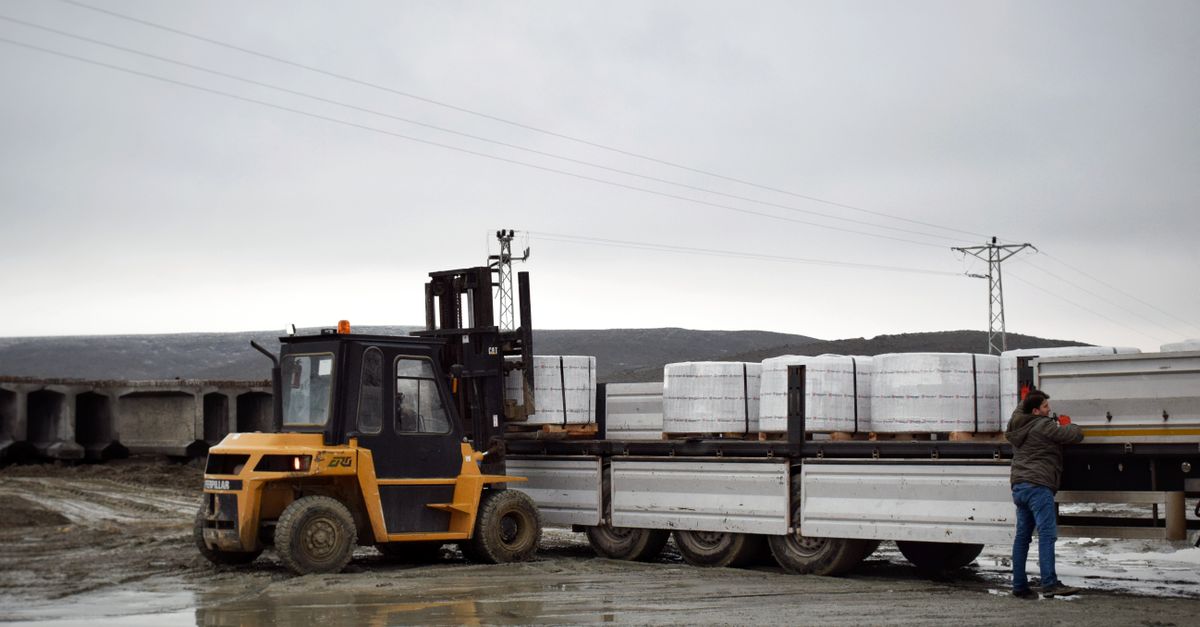 A forklift is loading cargo onto a truck in a muddy logistics area with a worker assisting.